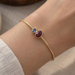 Könnte beinhalten: Ein dünner Goldarmreif mit zwei ovalen, in Zargenfassung gefassten Edelsteinen. Ein Stein ist ein kleiner blauer Saphir, der größere Stein ist ein tiefroter Rubin. Das Armband hat eine glatte, polierte Oberfläche und ein schlichtes Metalldesign.