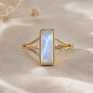Peut inclure: Une bague en or ornée d'une pierre de lune rectangulaire verticale présentant un éclat irisé bleu. L'anneau est fin et se divise en deux branches délicates qui se rejoignent sur les côtés du sertissage central, offrant un design équilibré.