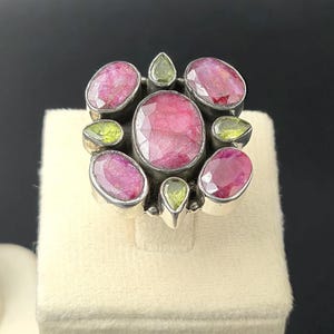 May include: A silver ring with a cluster of oval and pear-shaped gemstones. The ring features large, oval-cut pink rubies and smaller, pear-shaped green peridots. The gemstones are set in a silver band.