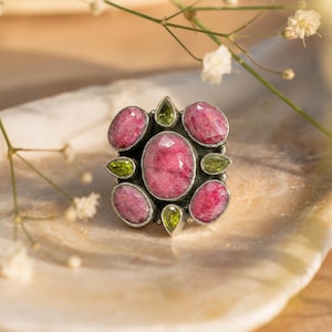 May include: A silver ring featuring a large central oval faceted pink stone surrounded by four smaller oval pink stones and four tiny pear-shaped green stones. The setting has a dark oxidized finish, creating a contrast with the vibrant pink and green gemstones.