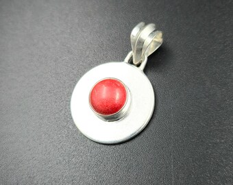 Puede incluir: Colgante de plata con diseño circular. El colgante presenta una piedra roja brillante, circular, engastada en el centro de un disco plateado. La fianza tiene un diseño decorativo y acanalado. El colgante se muestra sobre un fondo oscuro.
