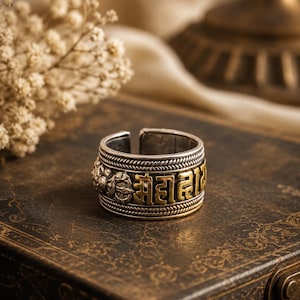Peut inclure: Une bague ouverte de couleur argentée avec une rangée centrale de caractères en écriture devanagari dorés en relief. La bague est bordée de détails en forme de corde torsadée et présente des motifs floraux ou décoratifs sur les côtés.