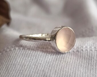 Puede incluir: Un anillo de plata con una gema grande, facetada, de color rosa pálido. La banda del anillo tiene un aspecto texturizado y martillado. La gema es redonda y está engastada en un engaste sencillo. El anillo se muestra sobre tela blanca.