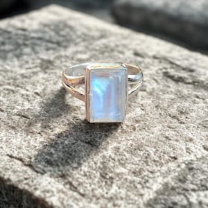 May include: A silver ring featuring a rectangular, iridescent, light blue gemstone. The ring's band splits into two parallel lines on each side of the gemstone. The gemstone has a subtle, glowing effect. The ring is set on a stone surface.