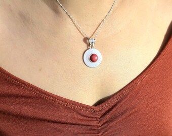 Puede incluir: Un collar con colgante de plata con un colgante circular y una piedra roja en el centro. El collar lo lleva una persona con una camiseta de color óxido. La cadena es delicada y el colgante es sencillo y elegante.