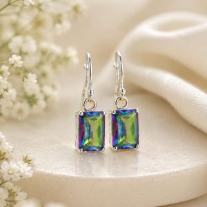 May include: A pair of silver drop earrings featuring rectangular-cut mystic topaz stones. The stones display a vibrant, iridescent spectrum of green, purple, and blue hues. Each earring is suspended from a simple silver hook wire setting.