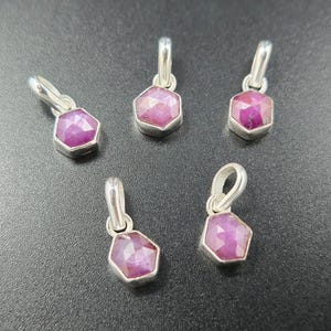 Puede incluir: Cinco colgantes de piedras preciosas de rubí con engastes plateados. Cada colgante presenta una piedra de rubí facetada y hexagonal. Los colgantes se muestran sobre un fondo oscuro, mostrando su vibrante color rosa-rojo y superficies reflectantes.