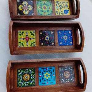 May include: Three wooden serving trays with decorative ceramic tile inserts. Each tray has a rectangular shape with curved handles. The tiles feature colourful floral patterns in yellow, green, blue, and other hues.