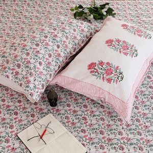 Könnte beinhalten: Ein Bettwäscheset mit einem weißen Laken und Kissenbezug mit dichtem rosa-türkisfarbenem Blumenrankenmuster, dazu ein zweiter weißer Kissenbezug mit lockerem rosa Blumenmotiv und rosa gestreiften Rändern sowie ein Notizbuch und ein Stift.