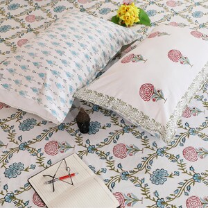 Può includere: Due cuscini rettangolari e un lenzuolo abbinato con base bianca e un motivo floreale ripetuto in blu, verde e rosso. Una piccola statuetta nera, un mazzetto di fiori gialli e un taccuino aperto con una penna rossa sono posati sopra.