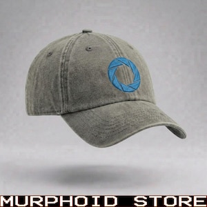 Puede incluir: Una gorra de béisbol gris carbón con una textura de tela desgastada y lavada. El panel frontal presenta un logotipo circular de apertura bordado en hilo azul vibrante, compuesto por ocho segmentos dispuestos en un patrón giratorio de obturador.