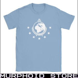May include: A light blue short-sleeve t-shirt featuring a white graphic print on the chest. The design displays a circular globe emblem surrounded by seven five-pointed stars, with a stylized rope-like border encircling the lower half of the globe.