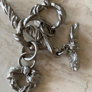 Könnte beinhalten: Halskette aus silberfarbenem Metall mit Knebelverschluss. Die Halskette hat ein verdrehtes Seildesign und einen herzförmigen Anhänger mit einer Schleife. Der Verschluss hat ein detailliertes, geformtes Design.