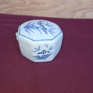Puede incluir: Un recipiente de cerámica blanco, octogonal, con diseños florales azules. La tapa y los lados presentan ilustraciones botánicas azules pintadas a mano. El recipiente tiene una pequeña asa curva en un lado. El fondo es una superficie burdeos y una superficie de madera marrón claro.