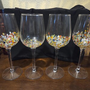 May include: Four clear wine glasses with colorful, speckled designs at the base. The glasses have long stems and rounded bowls. The speckled design includes yellow, blue, red, and white dots. The glasses are on a wooden surface.
