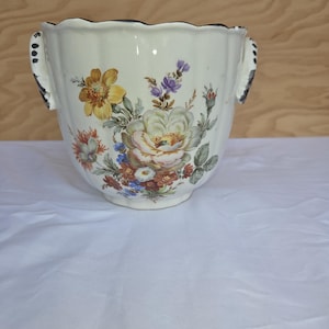 Może przedstawiać: Kremowy ceramiczny pojemnik na kwiaty z falistym brzegiem i dwoma uchwytami. Pojemnik zdobi kolorowy kwiatowy wzór z żółtymi, fioletowymi i czerwonymi kwiatami. Uchwyty mają czarne akcenty.