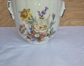 Puede incluir: Jardinera de cerámica color crema con borde festoneado y dos asas. La jardinera está adornada con un colorido diseño floral con flores amarillas, moradas y rojas. Las asas tienen detalles negros.