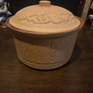 May include: A round, light brown ceramic casserole dish with a lid. The lid features a raised floral design. The dish has a textured, wood-like pattern on the sides. The lid has a small, round handle.