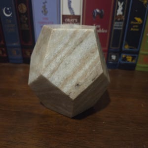 Puede incluir: Un objeto decorativo geométrico de múltiples lados hecho de piedra de color claro. La piedra tiene un aspecto natural veteado con vetas de color beige y crema. El objeto se exhibe sobre una superficie de madera oscura, con libros antiguos de fondo.