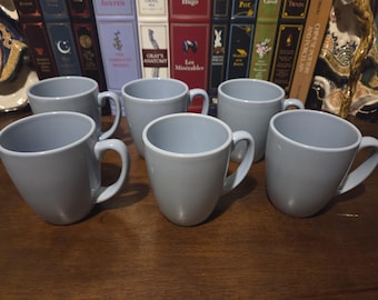 May include: Six light blue ceramic mugs with curved handles are arranged on a wooden surface. The mugs have a simple, modern design and a smooth, glossy finish. The background features a collection of books.