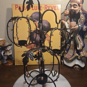 May include: A black metal candle holder featuring three pumpkin-shaped frames adorned with small amber-coloured beads and decorative metal leaves. The structure includes three circular bases for holding candles, supported by a central stand with curved legs.