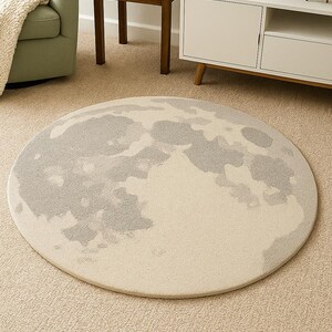 May include: A round rug with a moon design. The rug features a cream-colored background with a gray moon graphic. The rug is placed on a beige carpet in a living room setting.