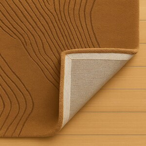 May include: A mustard-colored rug with a raised, linear pattern. The corner of the rug is folded back, revealing a beige backing with a woven texture and a cream-colored border. The rug is placed on a light wood surface.