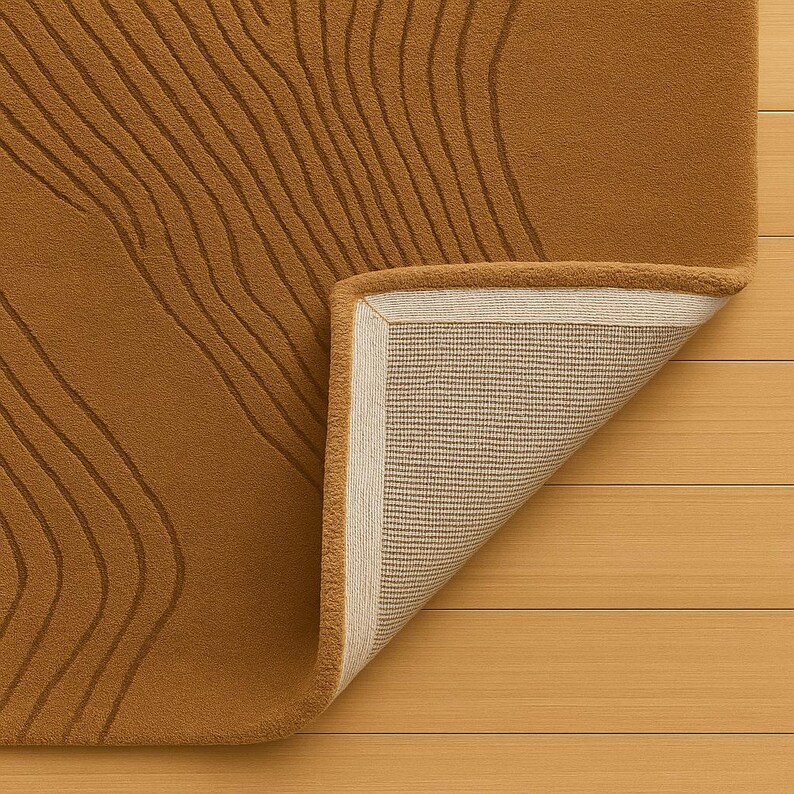 May include: A mustard-colored rug with a raised, linear pattern. The corner of the rug is folded back, revealing a beige backing with a woven texture and a cream-colored border. The rug is placed on a light wood surface.