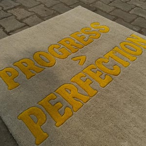 May include: A rectangular rug with the words "PROGRESS > PERFECTION" in yellow, bold, block letters. The rug has a neutral, light gray background. The rug is made of a soft, textured material.
