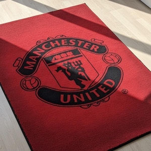 May include: A rectangular red doormat featuring the Manchester United logo in black. The logo includes the words "Manchester United" and a crest with a devil figure. The mat is designed for indoor use.