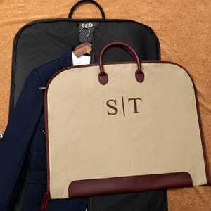 May include: A beige garment bag with dark red trim and handles, featuring the embroidered letters S|T in the center. The bag is positioned in front of a black garment bag containing a navy blue blazer and a white shirt on a wooden hanger.