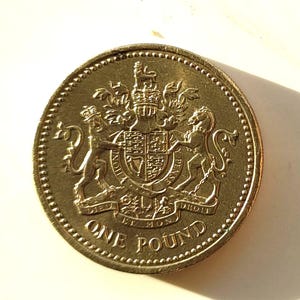 May include: A gold-coloured one-pound coin, featuring the Royal Coat of Arms. The coin shows a lion and unicorn supporting a shield, with the words "ONE POUND" inscribed on it.