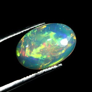May include: An oval-shaped opal gemstone featuring a translucent body with vibrant flashes of neon green, yellow, and orange play-of-color. The gemstone is held by a pair of silver-toned metal tweezers against a solid black background.