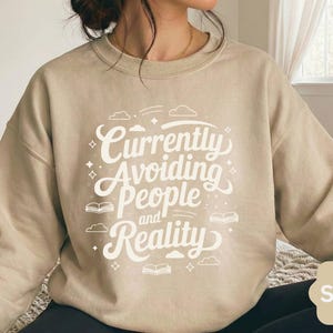 Peut inclure: Un sweat-shirt à col rond couleur sable avec un texte imprimé en blanc qui indique Currently Avoiding People and Reality. Le motif est agrémenté de petites illustrations graphiques de livres ouverts, de nuages et d'étoiles à quatre branches.