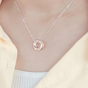 Peut inclure: Un collier argenté avec un pendentif composé de trois anneaux entrelacés en argent et or rose. Le collier est porté par une personne, avec la chaîne visible sur la peau et les vêtements.