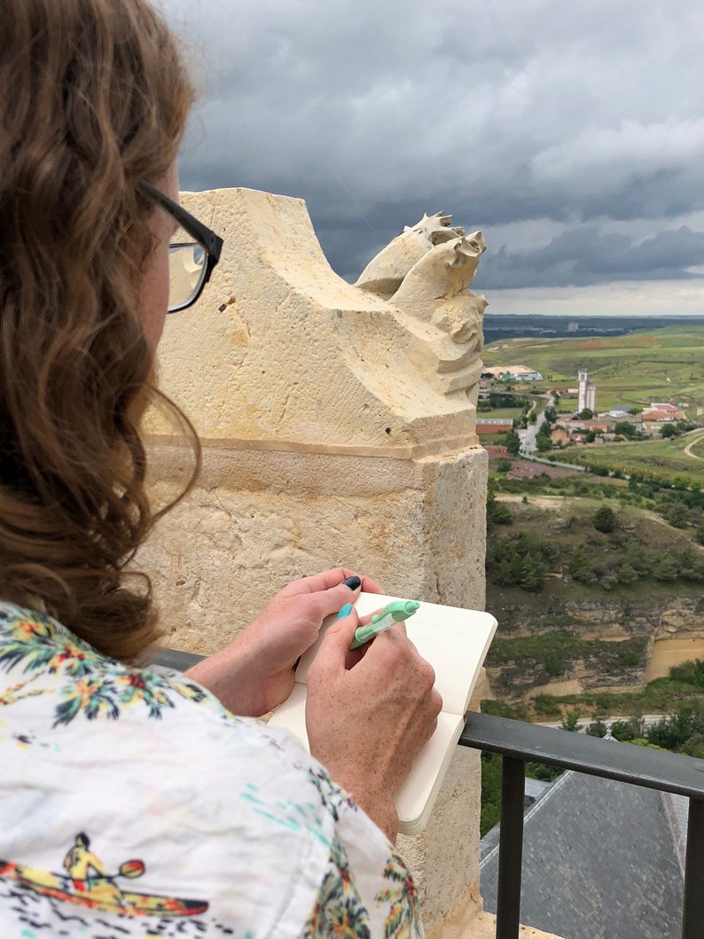 May include: A person is writing in a small, white notebook with a green pen while looking out at a view of a town and countryside. The person is standing on a stone balcony with a stone railing.