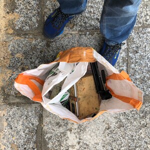 K&ouml;nnte beinhalten: Draufsicht auf eine wiederverwendbare Einkaufstasche, gef&uuml;llt mit Gegenst&auml;nden. Die Tasche ist wei&szlig; mit orangefarbenen Griffen und einem braunen Oberteil. Im Inneren befinden sich verschiedene Gegenst&auml;nde, darunter eine Holzkiste, ein schwarzes Stativ und andere Objekte. Blaue Turnschuhe und Jeans sind sichtbar.