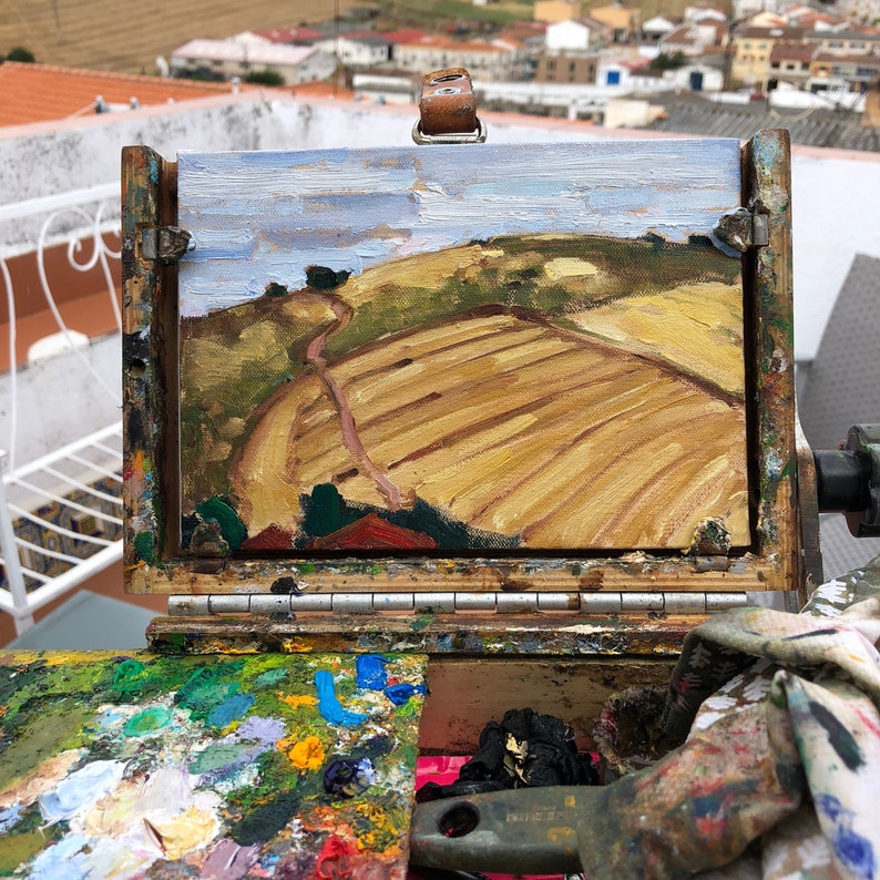 May include: A close-up of a small painting on an easel. The painting depicts a landscape with a field of yellow crops and a winding path leading up a hill. The sky is blue with white clouds.