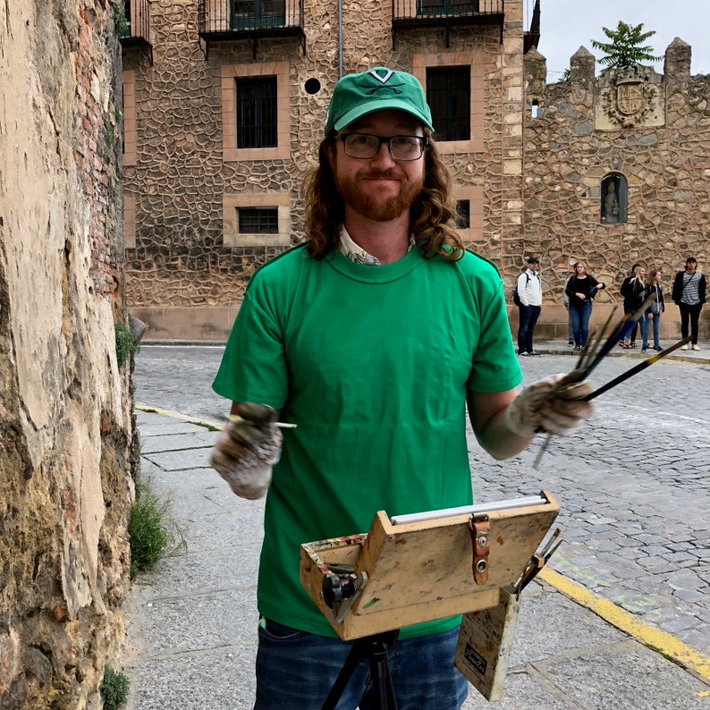 Puede incluir: Una persona con una camiseta verde y una gorra verde con un emblema blanco, sosteniendo pinceles y una paleta de artista de madera. El artista est&aacute; de pie en una calle adoquinada frente a un edificio de piedra.