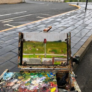 May include: A wooden easel with a canvas panel showing a painting of a landscape with a red traffic light. The easel has a paint palette with various colors of paint.