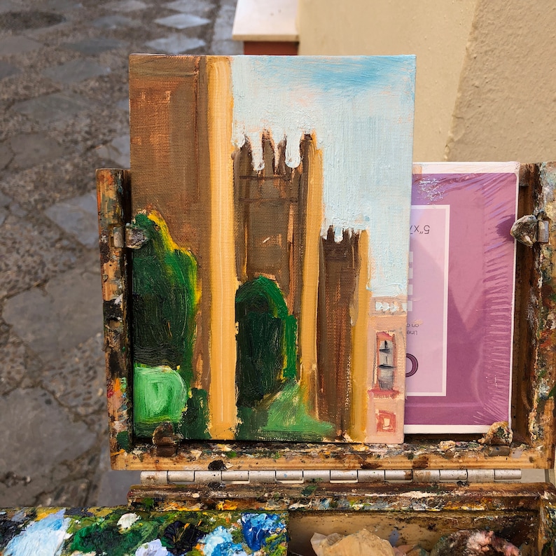 May include: An oil painting on canvas depicting a castle scene with brown and tan walls, green foliage, and a light blue sky. The painting is displayed in a wooden artist's box, with a pad of paper visible.