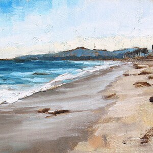 May include: Oil painting of a beach scene with a blue ocean and sky. The sandy beach has brown patches and the water has white waves. In the distance are mountains and trees. The artwork is painted with visible brushstrokes.