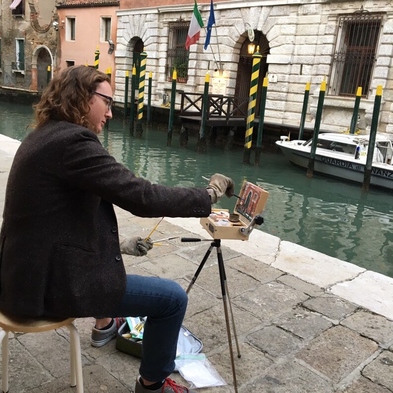 K&ouml;nnte beinhalten: Ein K&uuml;nstler malt im Freien, auf einem Hocker sitzend. Der K&uuml;nstler tr&auml;gt eine dunkle Jacke und blaue Jeans und arbeitet an einem kleinen Gem&auml;lde, das auf einem Stativ aufgebaut ist. Der Hintergrund zeigt einen Kanal mit Geb&auml;uden und einem Boot.