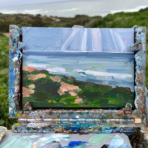 May include: An oil painting of a coastal landscape, featuring a view of the ocean under a cloudy sky. The painting is displayed on a weathered wooden easel, with a palette of paint colors visible below. The artwork captures the natural beauty of the scene.