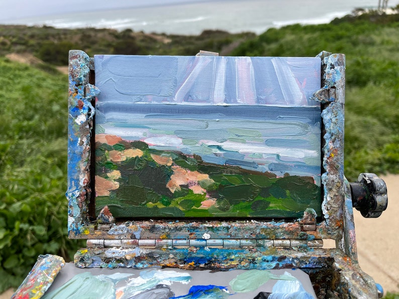 May include: An oil painting of a coastal landscape, featuring a view of the ocean under a cloudy sky. The painting is displayed on a weathered wooden easel, with a palette of paint colors visible below. The artwork captures the natural beauty of the scene.