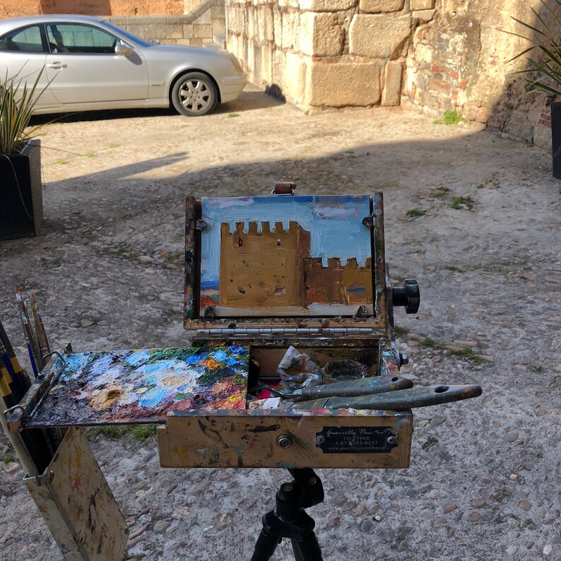 May include: An open wooden artist's paint box on a tripod, with a small painting of a castle. The palette is filled with various colors of paint. Paint knives and brushes are visible. The painting depicts a castle with a blue sky.