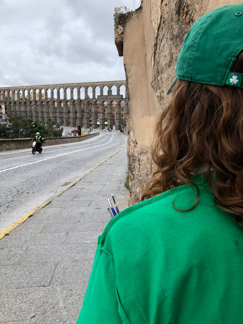 K&ouml;nnte beinhalten: Eine Person mit einem gr&uuml;nen T-Shirt und einer gr&uuml;nen Kappe mit einem wei&szlig;en Kleeblatt-Aufn&auml;her steht auf einem B&uuml;rgersteig. Im Hintergrund ein historisches Aqu&auml;dukt und eine Stra&szlig;e mit einem Motorrad.
