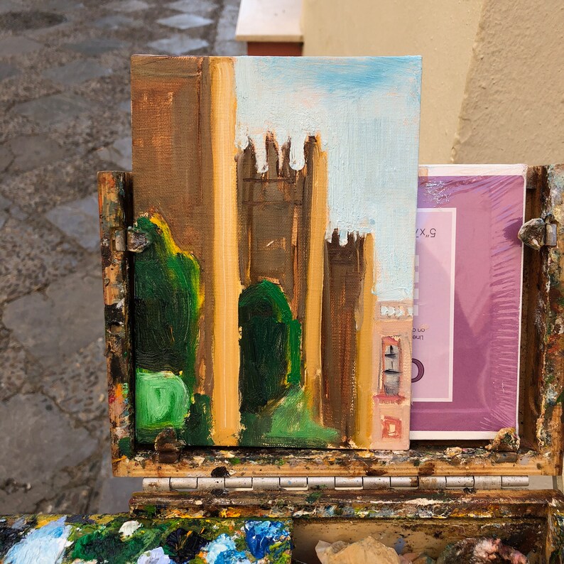 May include: A small painting on canvas depicting a brown building with a blue sky and green trees in the foreground. The painting is on an easel with a paint palette below it.