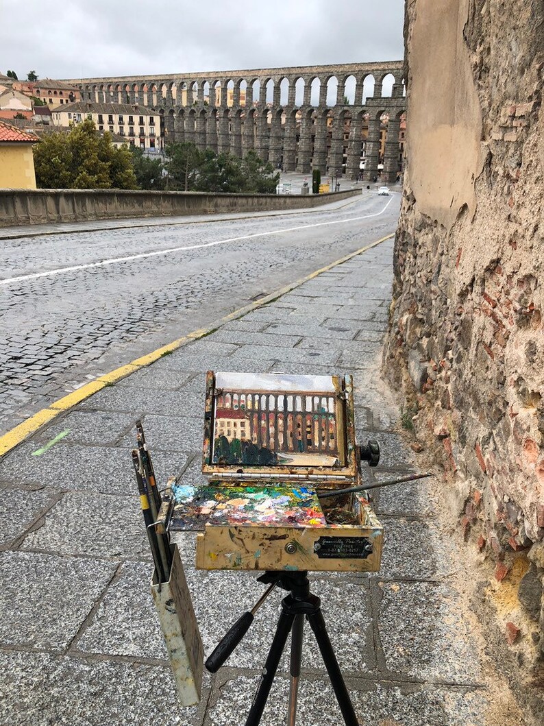 K&ouml;nnte beinhalten: Eine Staffelei mit einem Gem&auml;lde eines Aqu&auml;dukts, Pinseln und einer Palette mit bunten Farben. Die Staffelei steht auf einer Kopfsteinpflasterstra&szlig;e mit dem historischen Aqu&auml;dukt im Hintergrund. Die Szene wird an einem bew&ouml;lkten Tag aufgenommen.
