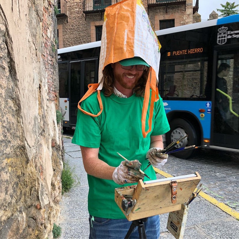 K&ouml;nnte beinhalten: Eine Person mit gr&uuml;nem T-Shirt, gr&uuml;ner Kappe und einer Einkaufstasche auf dem Kopf, die drau&szlig;en l&auml;chelnd malt. Sie h&auml;lt Pinsel und arbeitet an einer kleinen Staffelei. Im Hintergrund ist ein blauer Bus zu sehen.